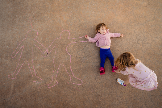 Two young children on pavement with chalk drawings of figures beside them.