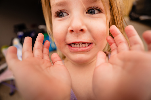 Young child with reddish-blonde hair smiling widely at camera with both hands raised toward lens.