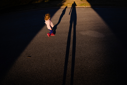 Child standing in sunlit street with elongated shadows of two adults cast across pavement.