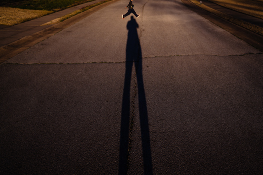 Person standing in the middle of an empty road with their elongated shadow stretching toward the camera.
