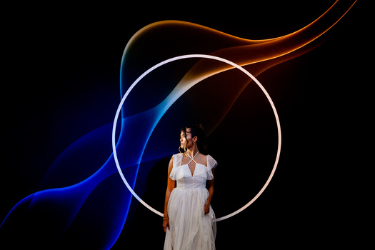 Woman in white dress standing within illuminated circular light rings against a dark background with blue and orange light trails.