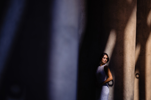 Woman in sunglasses between tall concrete pillars, standing in dramatic side-lit shadow.