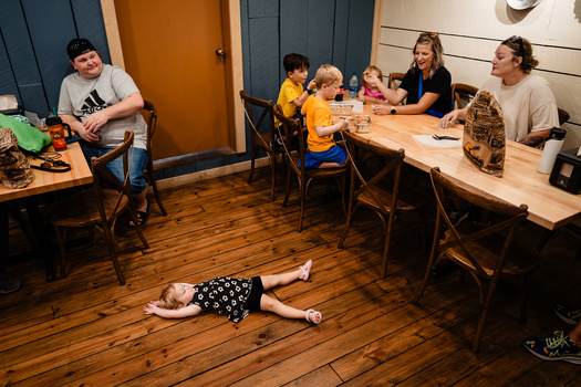Child lying on wooden floor while adults and children sit at tables during an indoor gathering.