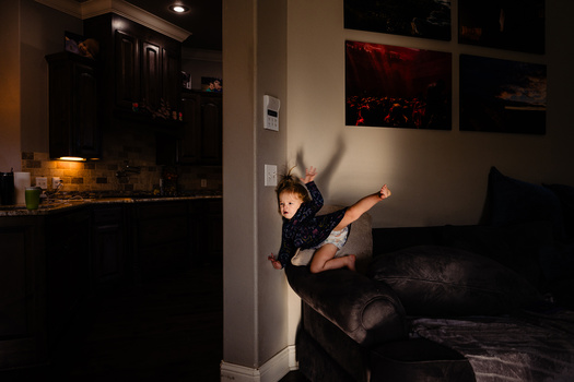 Child mid-jump on a dark couch in a dimly lit living room with warm accent lighting.