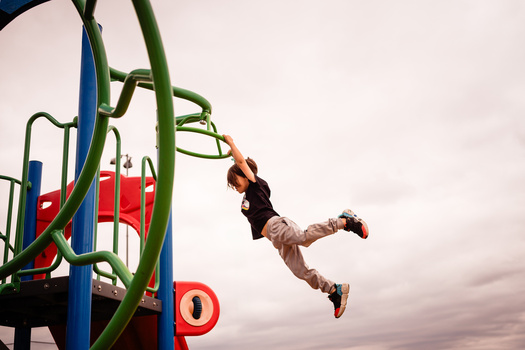 Child mid-jump reaching toward green playground equipment against overcast sky.