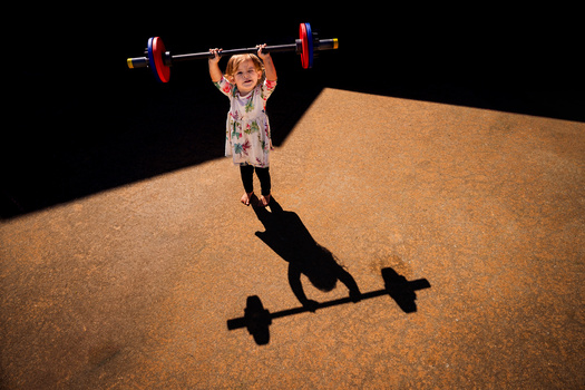 Child lifting a barbell on a sandy desert surface with dramatic shadow cast by studio lighting.