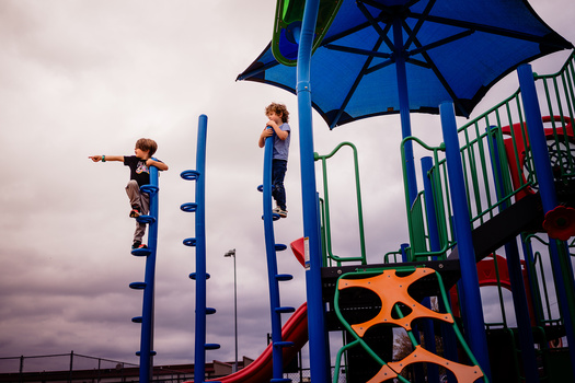 Children climbing on blue poles at a colorful playground structure beneath an umbrella canopy.