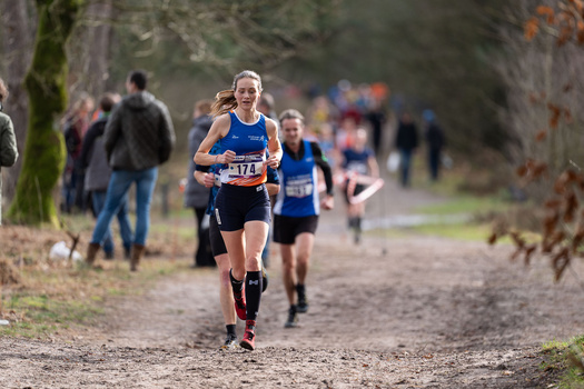 Female runner wearing blue race bib leads pack on tree-lined dirt path with spectators watching.