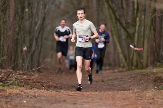 Trail runner in light-colored shirt and race bib leads a group of runners through a tree-lined forest path.
