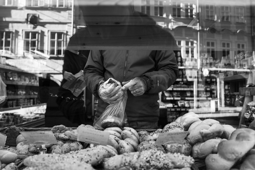 Vendor holding paper bag of vegetables at outdoor market stand with produce displayed below.