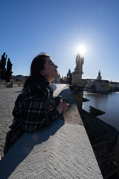 Woman sitting on a waterfront promenade with historic architecture and bright sunlight behind her.