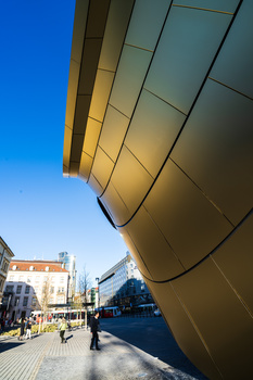 Curved metallic building facade with geometric panel divisions photographed from below against clear blue sky.