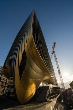 Dramatic upward view of a curved, metallic sculptural building with golden-bronze exterior panels against clear blue sky.