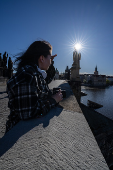 Photographer crouching on a riverside walkway, aiming a camera toward a sunlit cathedral and bridge.