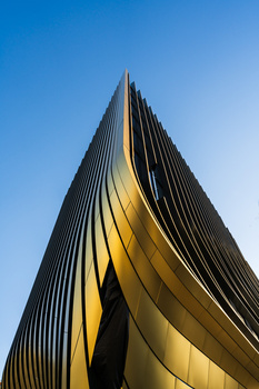 Modern skyscraper with curved gold and black striped facade shot from below against clear blue sky.