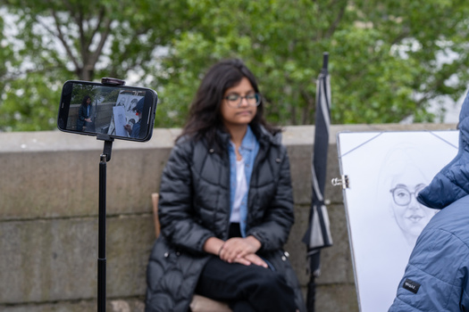 Woman in glasses and black jacket seated outdoors while being filmed on a smartphone mounted on a tripod.