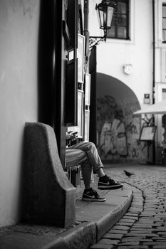 Person sitting alone on a curved bench against a white building wall in a narrow cobblestone alley.