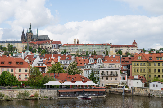 Historic riverside cityscape with terracotta-roofed buildings and Gothic spires rising above.