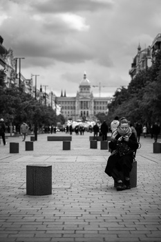 Woman in dark coat standing alone on wide plaza with classical government building in background under overcast sky.
