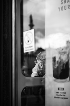 Black and white photograph of a child's face visible through a train or bus window, with blurred landscape reflections.
