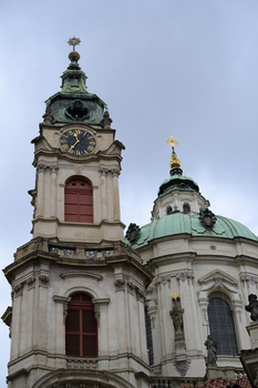 Baroque church tower with cream-colored facade, ornate clock face, and green copper dome beneath overcast sky.