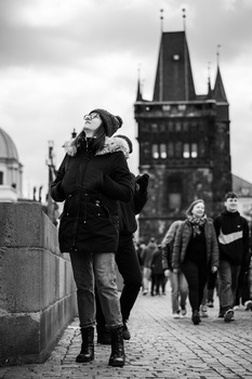 Woman in dark coat walking on cobblestone street with Gothic architecture in background.