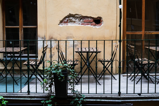 Weathered cream-colored facade with damaged stucco patch and iron balcony railing lined with folding chairs.