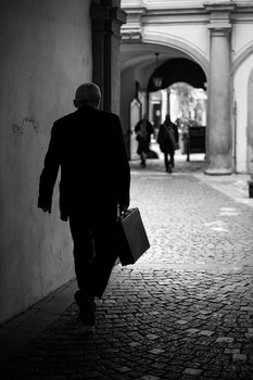 Man in dark suit walking away through a European-style covered arcade, carrying a briefcase.