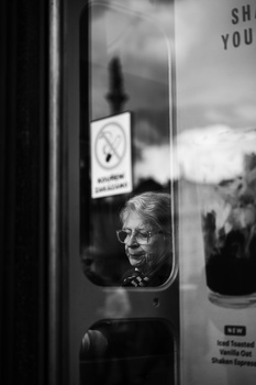 Elderly man with glasses visible through a glass door or window with reflections of clouds and landscape.