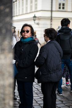 Woman in sunglasses and dark jacket stands on cobblestone street, engaging with another person.