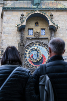 Two people viewing an ornate astronomical clock tower with blue decorative elements and an arched alcove.