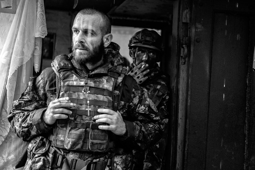 Two soldiers in camouflage uniforms and tactical gear stand in a doorway during rainfall.