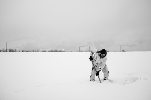 Person in white winter camouflage crouching in deep snow against a gray sky and distant treeline.