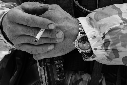 Close-up of two hands holding a cigarette, wearing formal attire and watches, photographed in black and white.