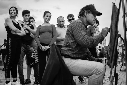 Man in plaid shirt and cap crouches while group watches in black and white photograph.