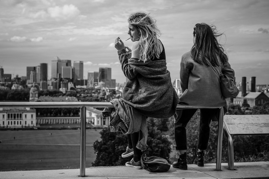 Two women sitting on a railing overlooking a city skyline in black and white.