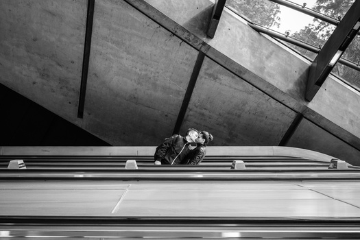 Person crouching on narrow metal beam suspended high above ground within brutalist concrete structure.