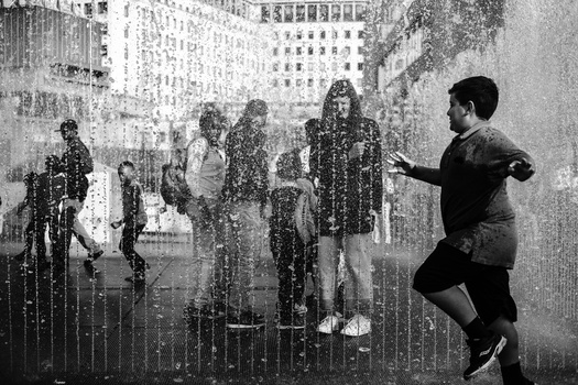 Black and white photograph of people playing in an urban water fountain with city buildings in the background.