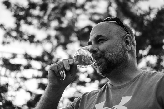 Bearded man drinking from a glass outdoors with blurred foliage in the background.