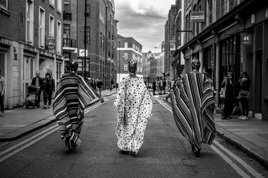 Three people wearing striped and patterned wraps walk down a busy urban street in black and white.