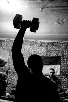 Silhouetted figure holding a dumbbell overhead against a cloudy sky in this high-contrast black and white photograph.