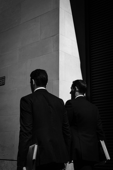 Two people in dark business suits photographed from behind against a minimalist architectural background.