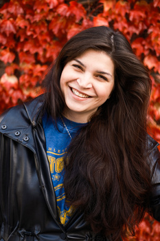 Smiling woman with long dark hair wearing a black leather jacket and blue graphic shirt against a red autumn foliage background.