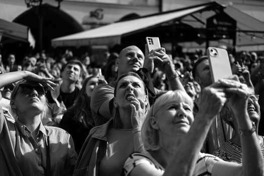 Crowd of people looking upward with phones and cameras raised, captured in black and white.