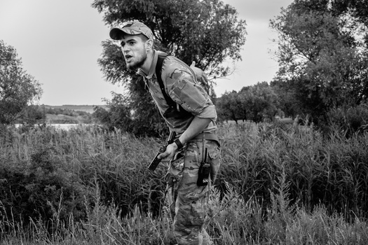 Man in camouflage clothing and cap crouching in overgrown field with trees in background.