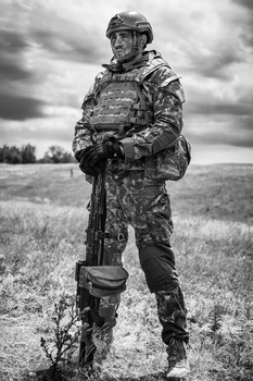 Soldier in camouflage uniform and helmet standing in an open field under overcast sky.