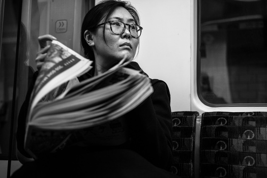 Young man wearing glasses on public transit, holding a newspaper, looking upward in black and white.