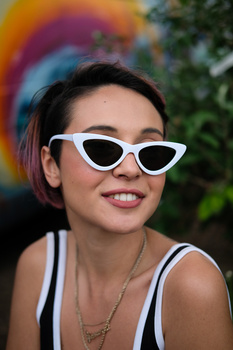 Young woman wearing white cat-eye sunglasses and a striped tank top, smiling at camera with colorful blurred background.