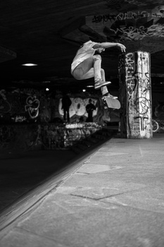 Skateboarder performing aerial trick in indoor skate park with spectators watching.