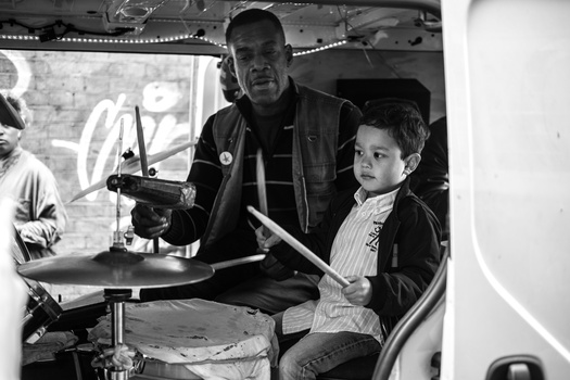 Man and young boy sit at drums in what appears to be a tour bus or vehicle interior.